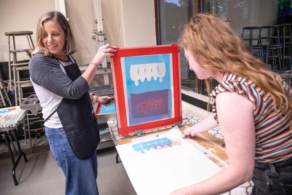 Two women are engaged in a screen printing session, with colorful designs displayed on a table in a creative studio environment.