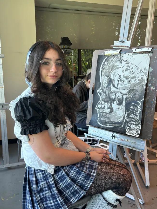 A seated artist in a black and white outfit poses next to an easel displaying a skeletal drawing in a bright, studio-like setting.
