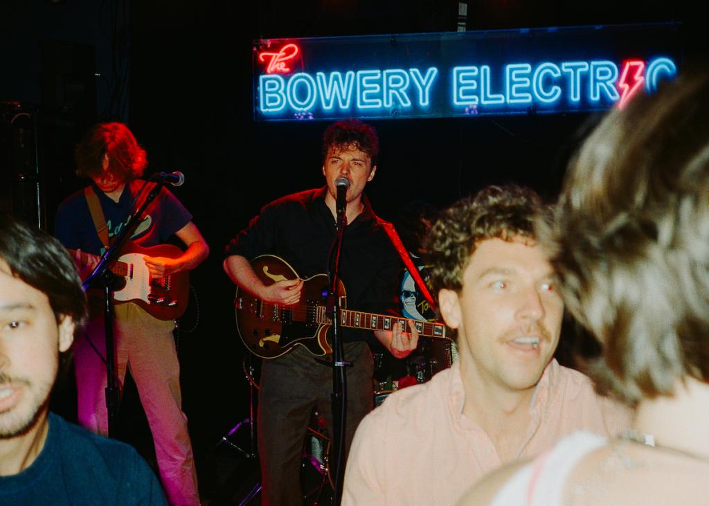 A lively performance at The Bowery Electric, featuring musicians playing guitar and drums, with an energetic audience in the foreground.
