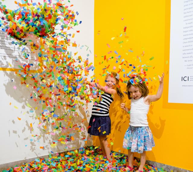 Two young visitors are throwing confetti into the air and standing under it as it falls.