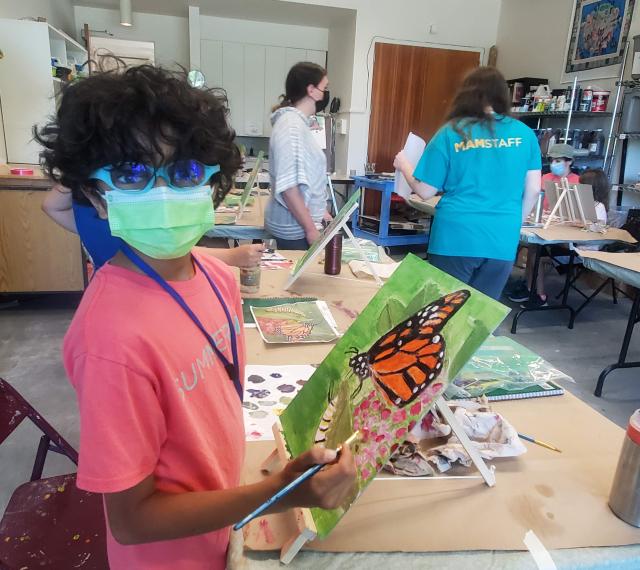 A young student is wearing a mask and looking at the camera while painting a work on canvas.