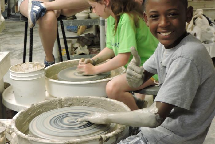A young student is smiling at the camera and sitting at a pottery wheel. He is covered in clay up to his elbows and looks pretty happy about it. 