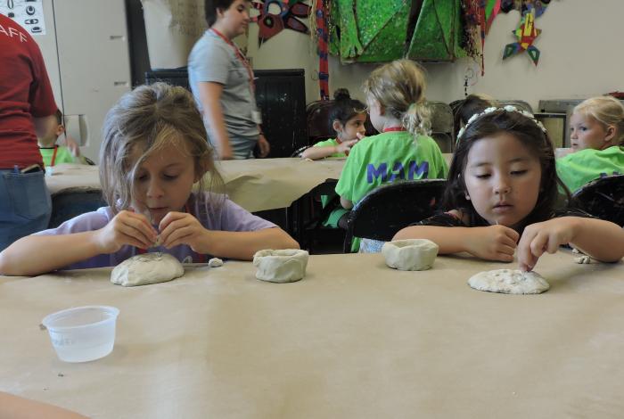 two children are working at a table next to each other on clay projects. They are both focused on their artwork and not looking at the camera.