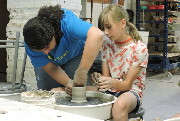 an instructor is showing a student how to make her piece taller on the pottery wheel. The student is watching. 