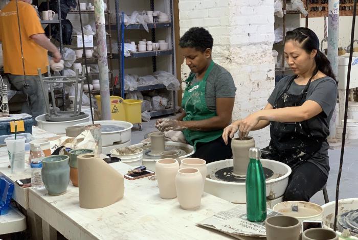 two students are working on the pottery wheels in the ceramic studio. Both are focused on their clay cylinders and not looking at the camera. Instructor Steve is in the background, looking a finished pieces on the shelves. 