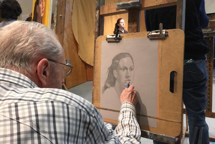 A student is sitting at an easel, drawing a portrait of a woman with a pencil.