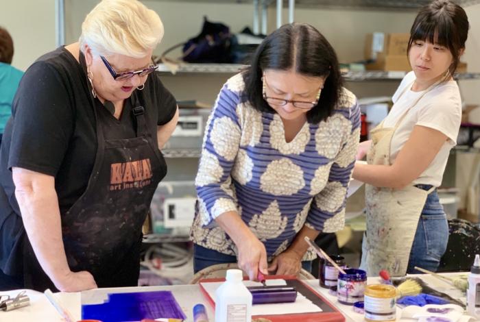 A woman is rolling ink onto a breyer with dorothy cochran standing next to her and looking on. Another student is behind them watching.