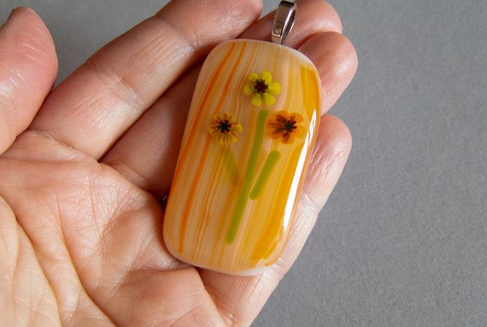 a close up picture of a hand holding a pendant made of fused glass. glass is in a floral pattern with an orange tonal striped background. 