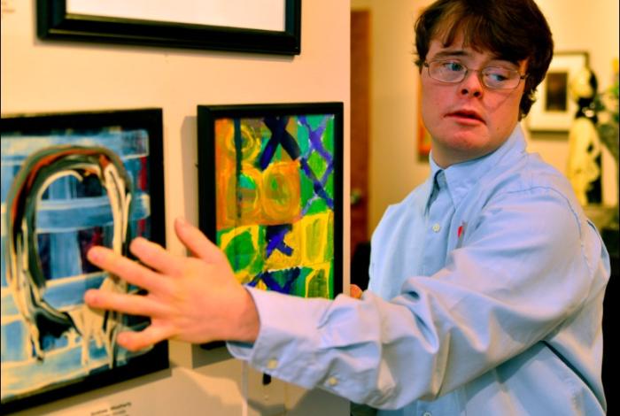 An artist is pointing to an artwork in the gallery on the left and looking to the right.