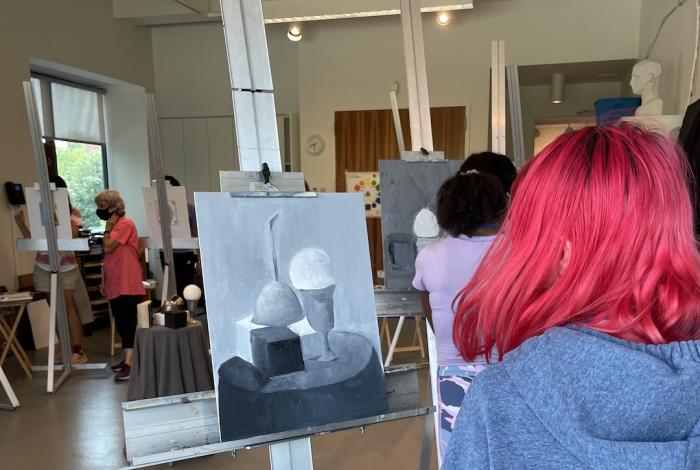 Looking over the should of a student with bright red hair at their greyscale still life painting in progress on an easel.