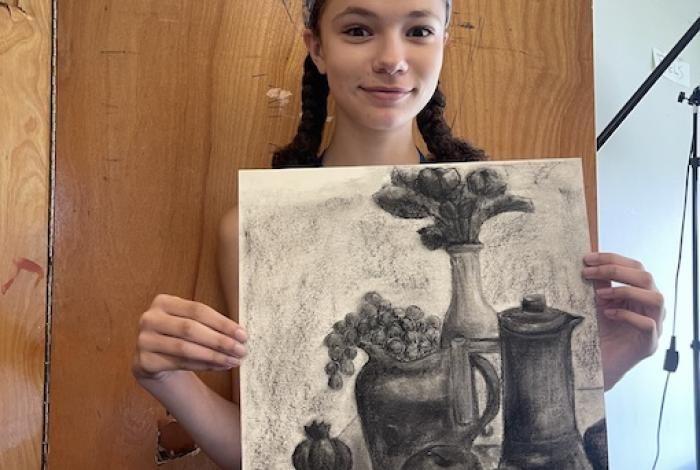 Teen student holding up a charcoal still life drawing containing a vase of flowers, a pitcher, grapes, an apple, and an orange