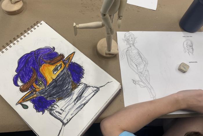 Sketches on paper, one with a colorful stylized character and another with a human figure, beside a wooden mannequin.