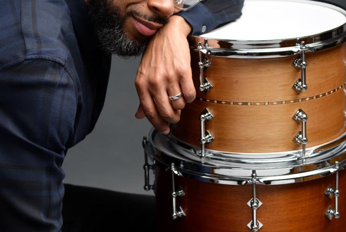 A person in a dark plaid shirt leans on a wooden drum, showcasing their hands adorned with rings. The background is softly blurred.