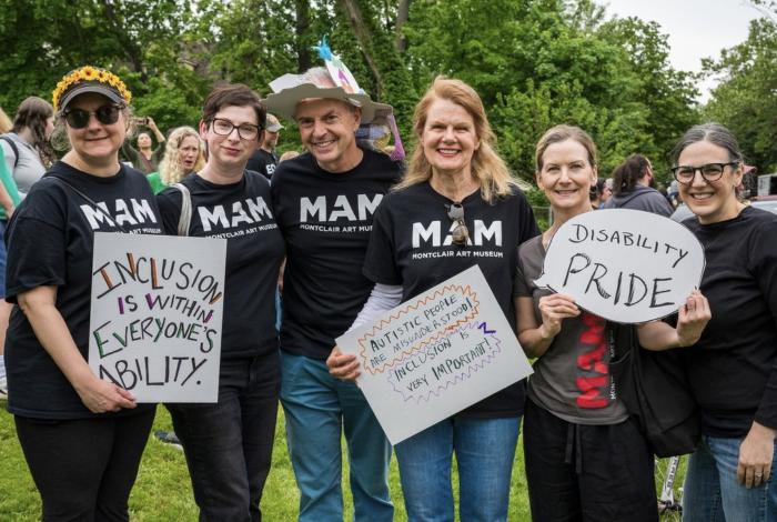 A group of individuals wearing "Montclair Art Museum" T-shirts holding signs advocating for inclusion and disability pride at an outdoor event.