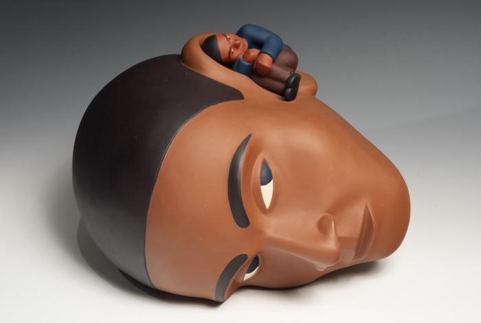 A sculptural figure showcases a stylized person reclining on a round, smooth surface, with warm earthy tones and distinct features.