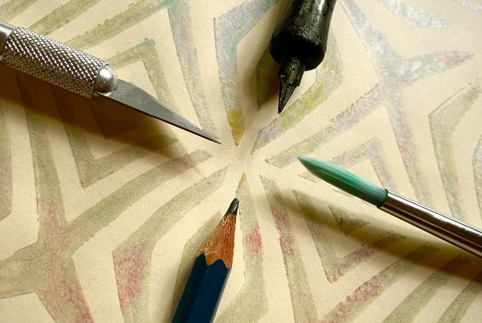 Art supplies arranged around a colorful patterned paper, featuring a craft knife, pencil, green paintbrush, and ink pen.