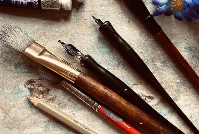 A variety of art supplies including brushes, pencils, paint tubes, and flowers arranged on a textured surface.