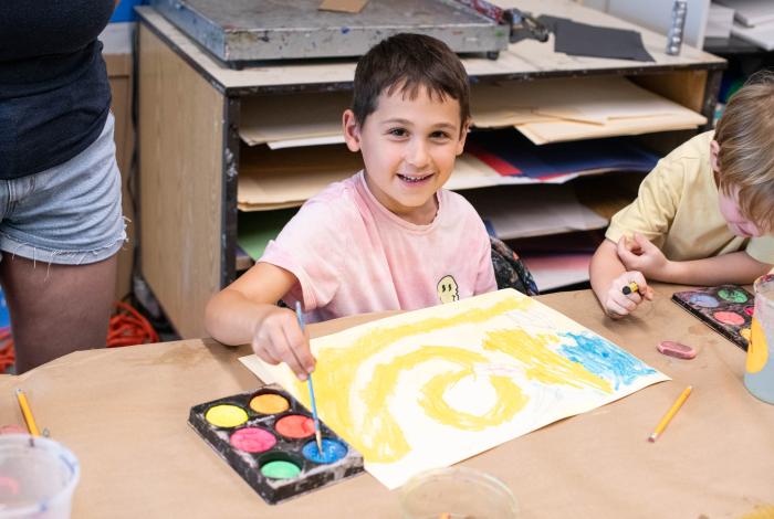 A child in a pink shirt paints a colorful swirl on paper, while another child observes, surrounded by art supplies in a classroom.