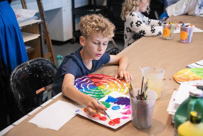 A young child paints a colorful rainbow design on paper at a table, surrounded by art supplies and other children engaged in creative activities.