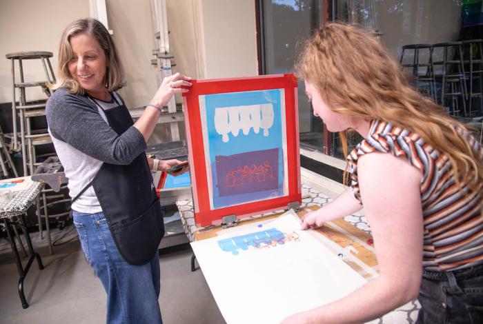 Two women are engaged in a screen printing session, with colorful designs displayed on a table in a creative studio environment.