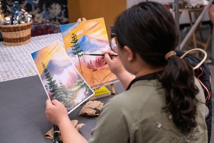 A woman paints on a canvas with a scenic mountain backdrop.