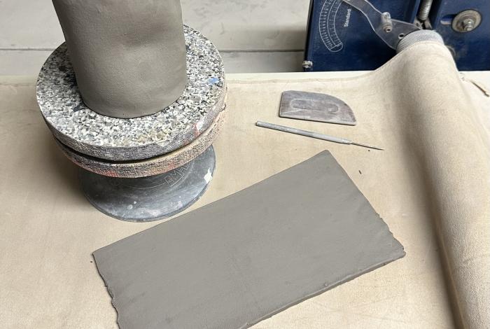 A workbench featuring a cylindrical clay piece on a stone base, a flat slab of clay, and various pottery tools.