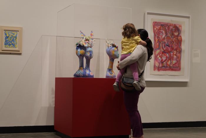 A mother holds her daughter while admiring colorful sculptures in a museum, with abstract art displayed on the walls.