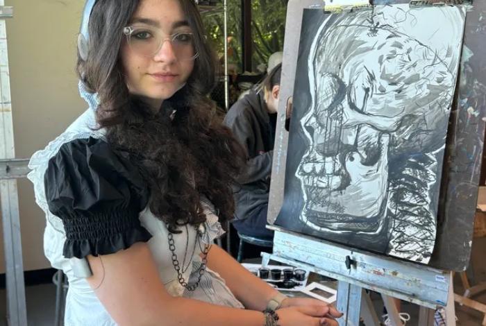 A seated artist in a black and white outfit poses next to an easel displaying a skeletal drawing in a bright, studio-like setting.