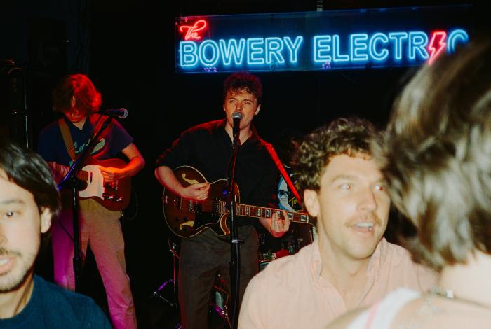 A lively performance at The Bowery Electric, featuring musicians playing guitar and drums, with an energetic audience in the foreground.