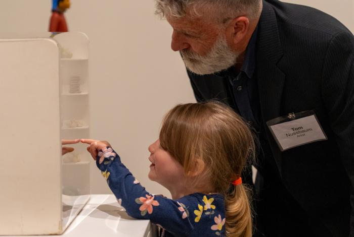 A child in a floral dress curiously points at an exhibit inside a gallery, while an adult leans in thoughtfully beside them.